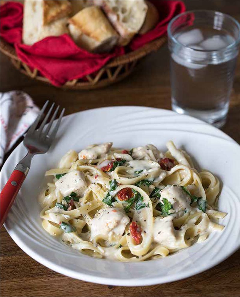 Tuscan Garlic Chicken from the Electric Pressure Cooker Cookbook - featuring 45 degree shot of fettuccini noodles with a white garlic cream sauce, parsley, sundried tomatoes, and chicken, with a red fork on the side