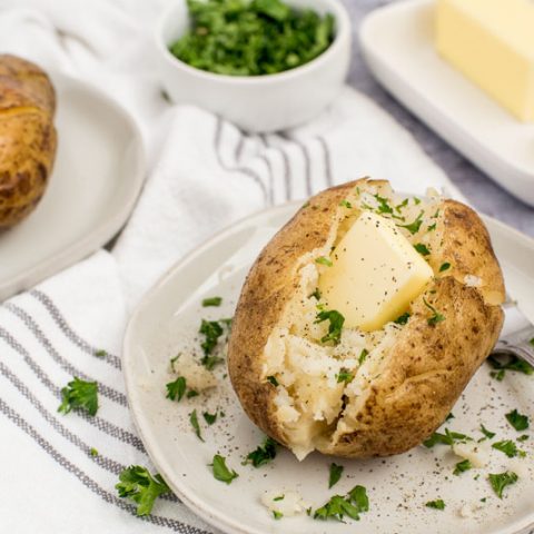 Instant pot baked potatoes with a pat of butter and fresh herbs sprinkled on top on a white plate with a butter dish in the background