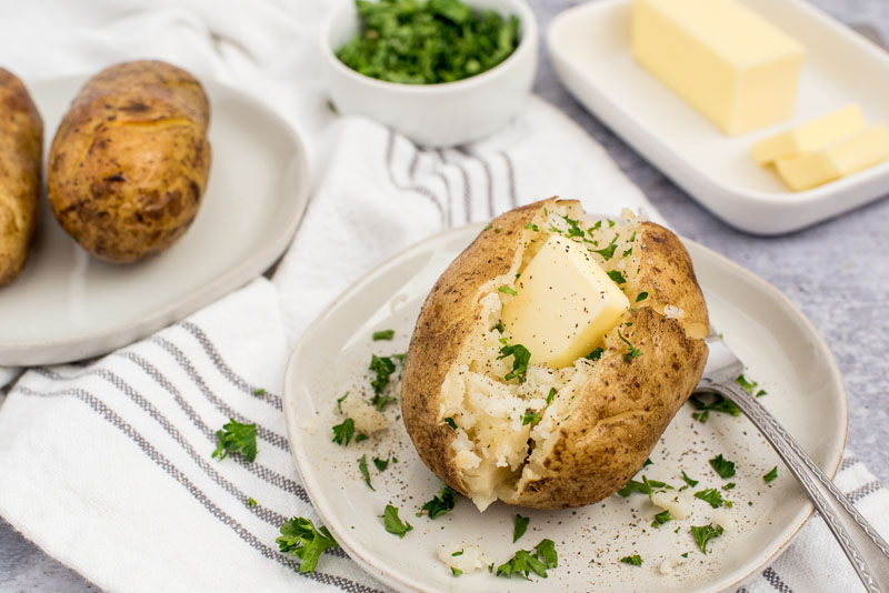 Instant pot baked potatoes with a pat of butter and fresh herbs sprinkled on top on a white plate with a butter dish in the background