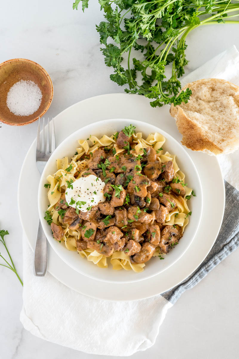 An overhead shot of a bowl of beef stroganoff served over egg noodles, with a roll parsley and a pinch bowl of salt in the background