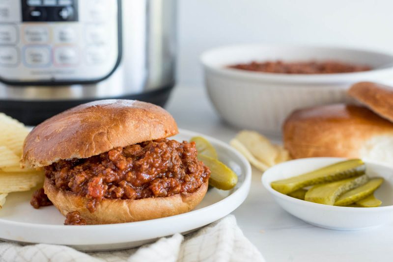 Sloppy Joe served on a bun with pickles, rolls, sloppy box mix, and an Instant Pot in the background.