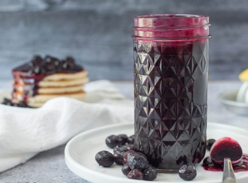 a textured mason jar filled with blueberry compote with extra blueberries on a white plate with a stack of pancakes topped with blueberry sauce in the background