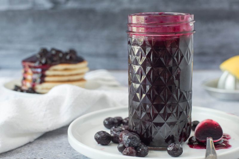 a textured mason jar filled with blueberry compote with extra blueberries on a white plate with a stack of pancakes topped with blueberry sauce in the background
