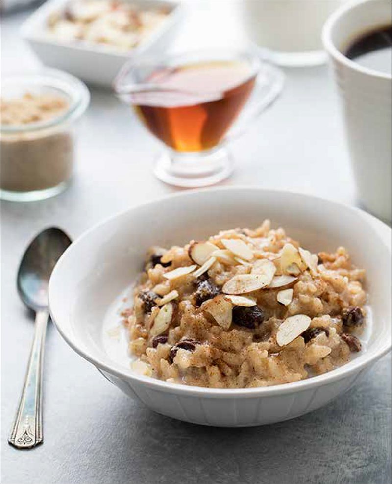 Maple Almond Risotto from the Electric Pressure Cooker Cookbook - featuring 45 degree shot of a white bowl filled with a tan risotto, a splash of milk, raisins, and sliced almonds with a dash of cinnamon on top