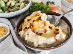 A 45 degree shot of a silver spoon dipping into a bowl of caramelized onion mashed potatoes, topped with shredded cheese and bacon