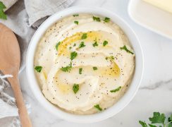 An overhead shot of a white bowl filled with cauliflower mashed potatoes, topped with butter, salt, and pepper