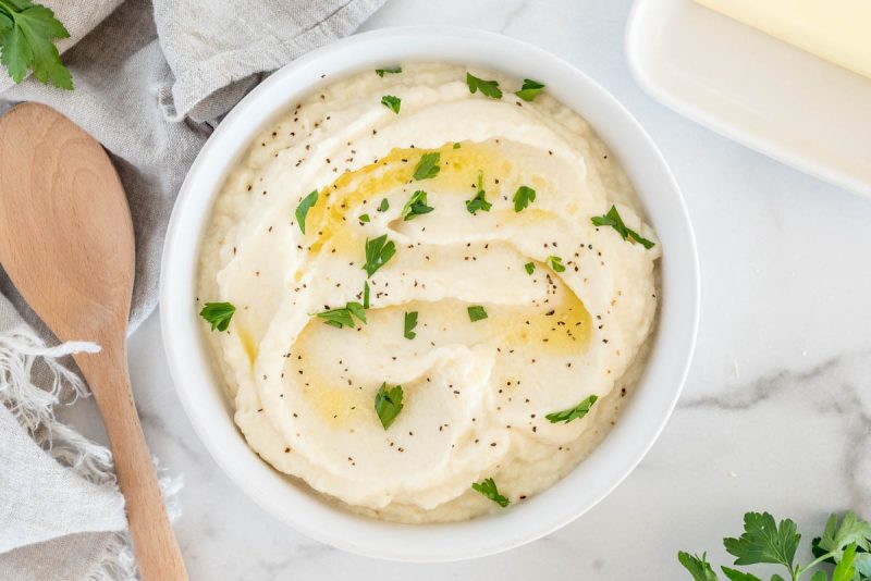 An overhead shot of a white bowl filled with cauliflower mashed potatoes, topped with butter, salt, and pepper