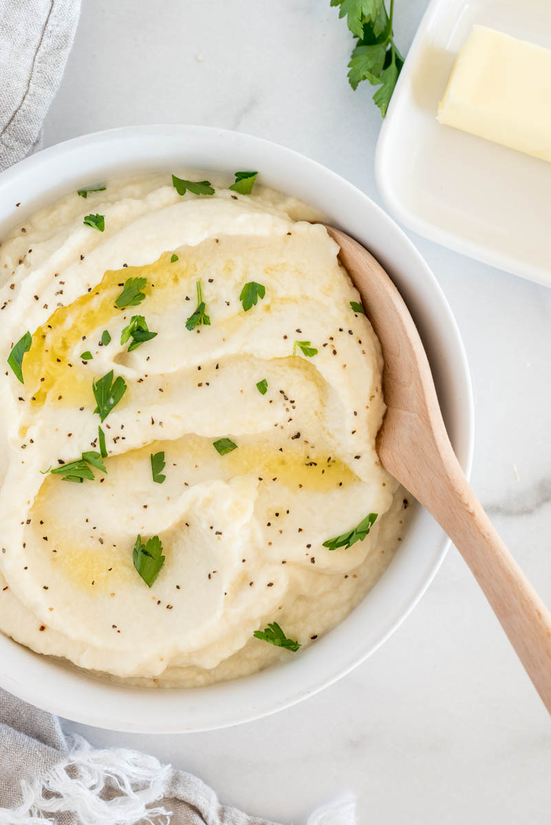 And overhead shot of a white bowl filled with mashed cauliflower, with a wooden spoon ready to serve