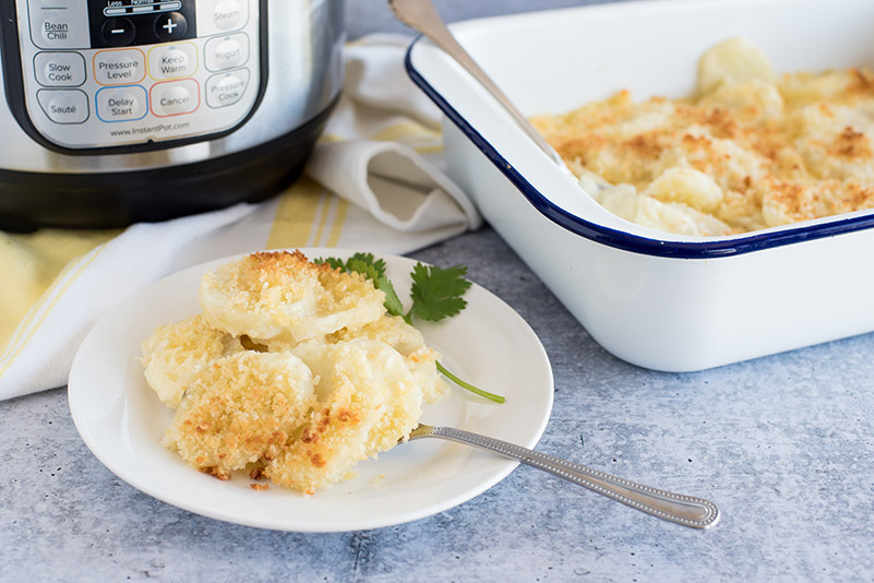 Instant Pot Scalloped Potatoes, plated on a small white plate with an Instant Pot Duo and a white baking dish in the background