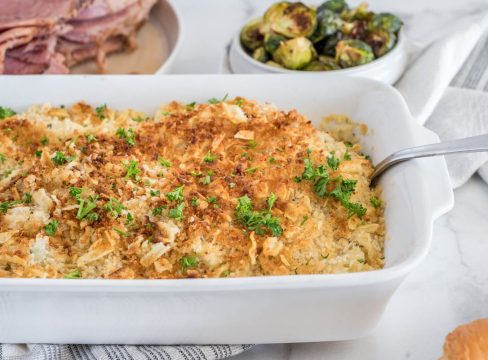 A serving dish with Instant Pot hash brown casserole and a serving spoon, placed in front of sliced ham and roasted Brussel sprouts.