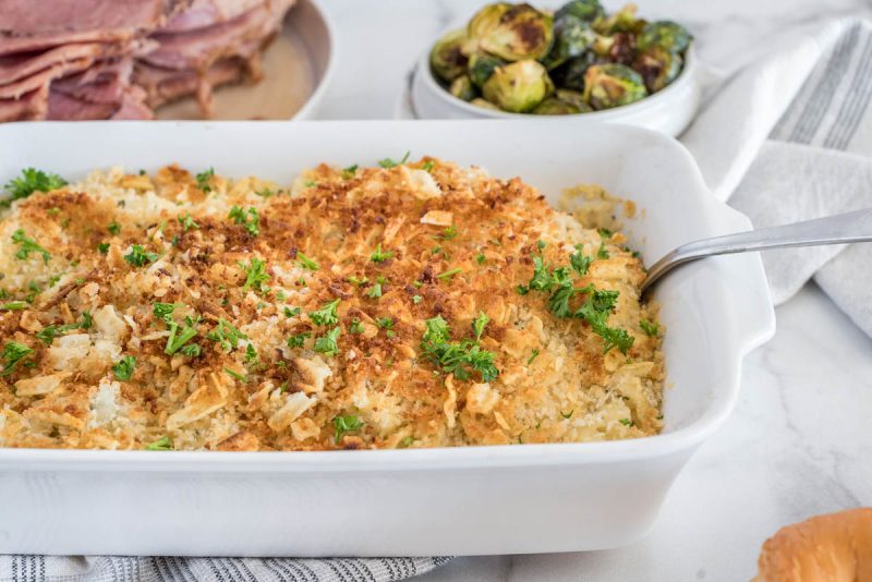 A serving dish with Instant Pot hash brown casserole and a serving spoon, placed in front of sliced ham and roasted Brussel sprouts.