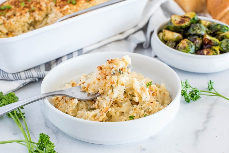 Instant Pot hash brown casserole served in a white dish with a fork with fresh parsley and roasted Brussel sprouts in the background.