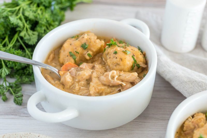 Close up picture of a white bowl with Instant Pot chicken and dumplings, with fresh parsley and another bowl of chicken and dumplings in the background.