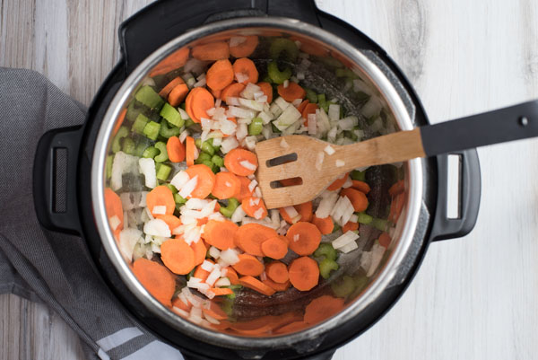 Overhead of Instant Pot filled with carrot rounds, diced onion and celery being sautéed with a wooden spoon to make chicken and wild rice soup.