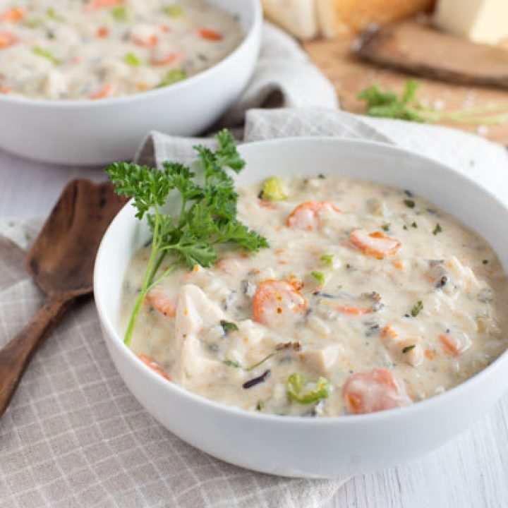 two white bowls filled with creamy Instant Pot chicken wild rice soup garnished with parsley on a grey checkered cloth napkin with a wooden spoon.