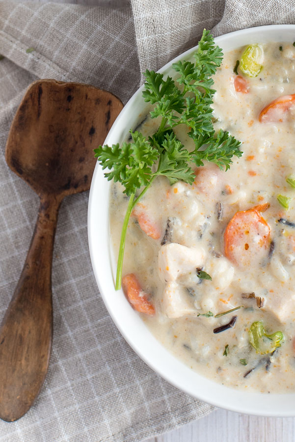 Overhead of white bowl of pressure cooker chicken and wild rice soup garnished with parsley served with crusty white bread and butter styled with a grey and white checkered cloth napkin, wooden spoon.