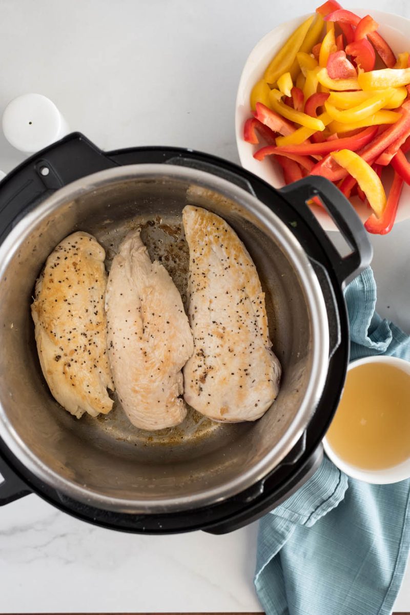 An overhead shot of chicken tenders browned in the Instant Pot with sliced bell peppers and broth in the background