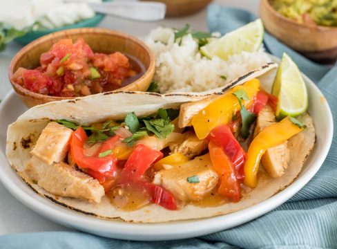 A side shot of a fajita, loaded with peppers, cilantro, and chicken, garnished with a lime with salsa and rice in the background