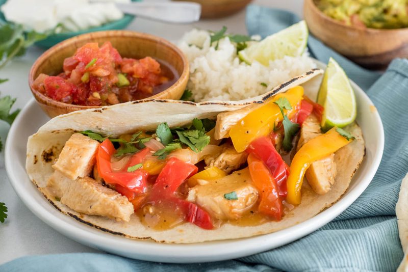 A side shot of a fajita, loaded with peppers, cilantro, and chicken, garnished with a lime with salsa and rice in the background