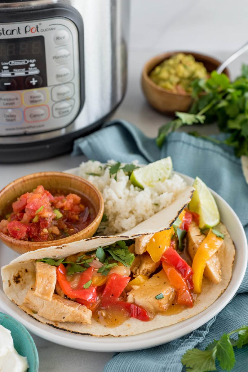 a 45 degree shot of a white plate with a chicken fajita filled with chicken, peppers, and cilantro, with a small bamboo bowl of salsa and lime rice in the background, with an Instant Pot and guacamole behind