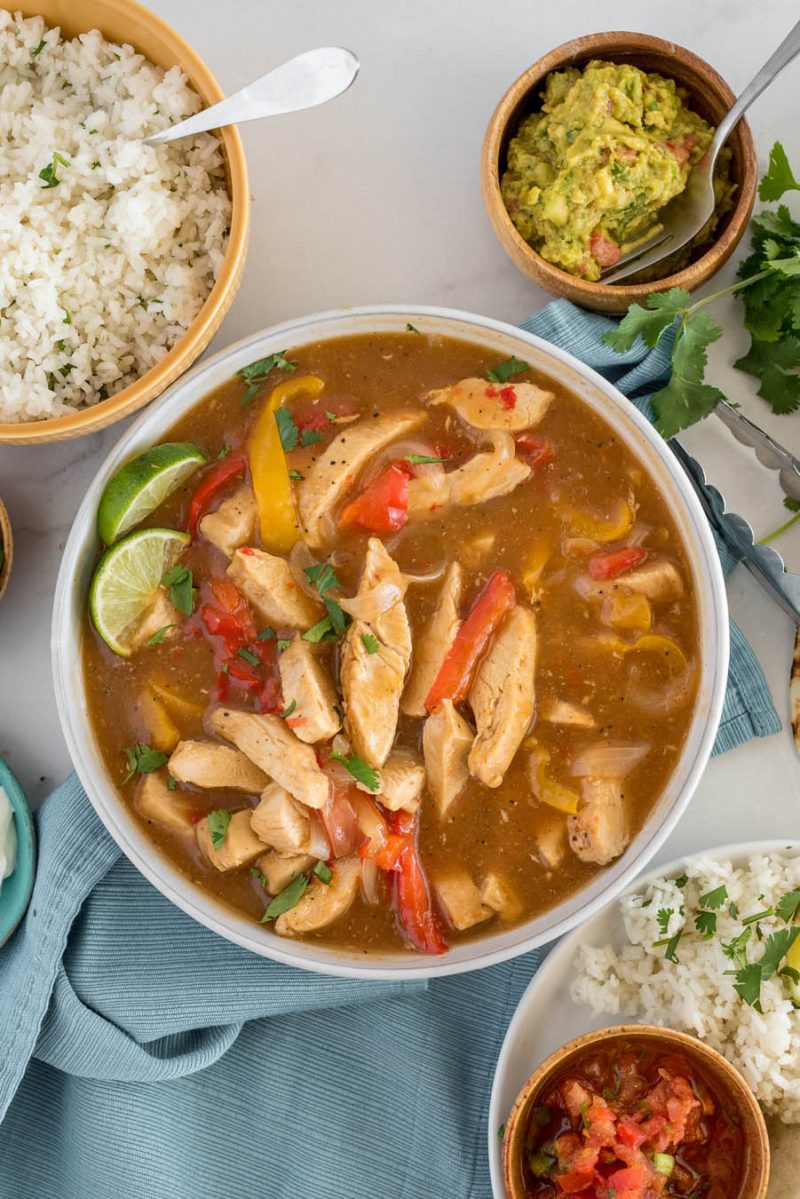 An overhead vertical shot of a bowl filled with fajita fixings—sliced chicken, red and yellow bell peppers, and onions garnished with cilantro and lime wedges, with a bowl of rice, guacamole, and salsa