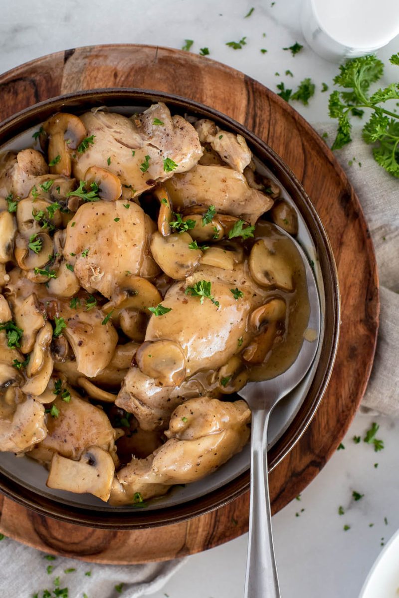 An overhead shot of the chicken marsala with a silver spoon, garnished by parsley on a wooden round serving plate