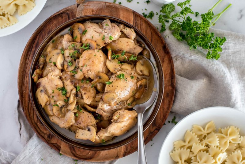 an overhead shot looking into a dark bowl on a wooden platter, filled with cooked chicken marsala ready to spoon onto pasta