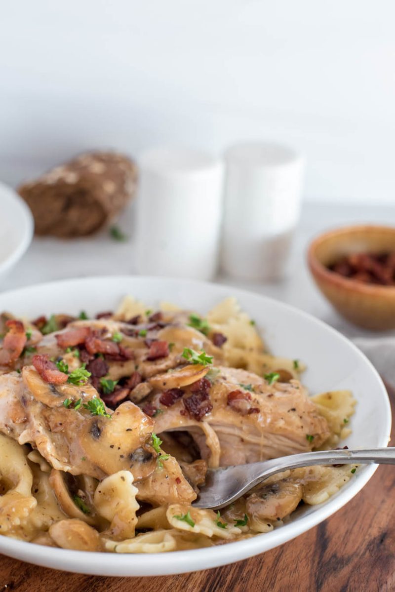 A vertical 45 degree shot of a white bowl filled with chicken marsala, garnished with bacon and parsley, served over a bed of yellow bowtie pasta