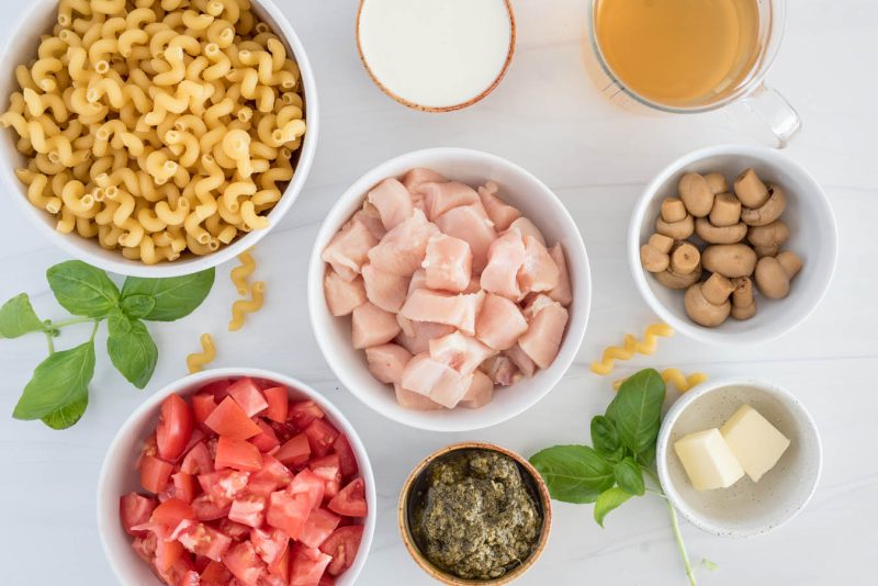 Overhead shot of all the ingredients for Instant Pot chicken pesto pasta, including cavatapi noodle, diced chicken, diced tomatoes, pesto, butter, mushrooms, and chicken broth.