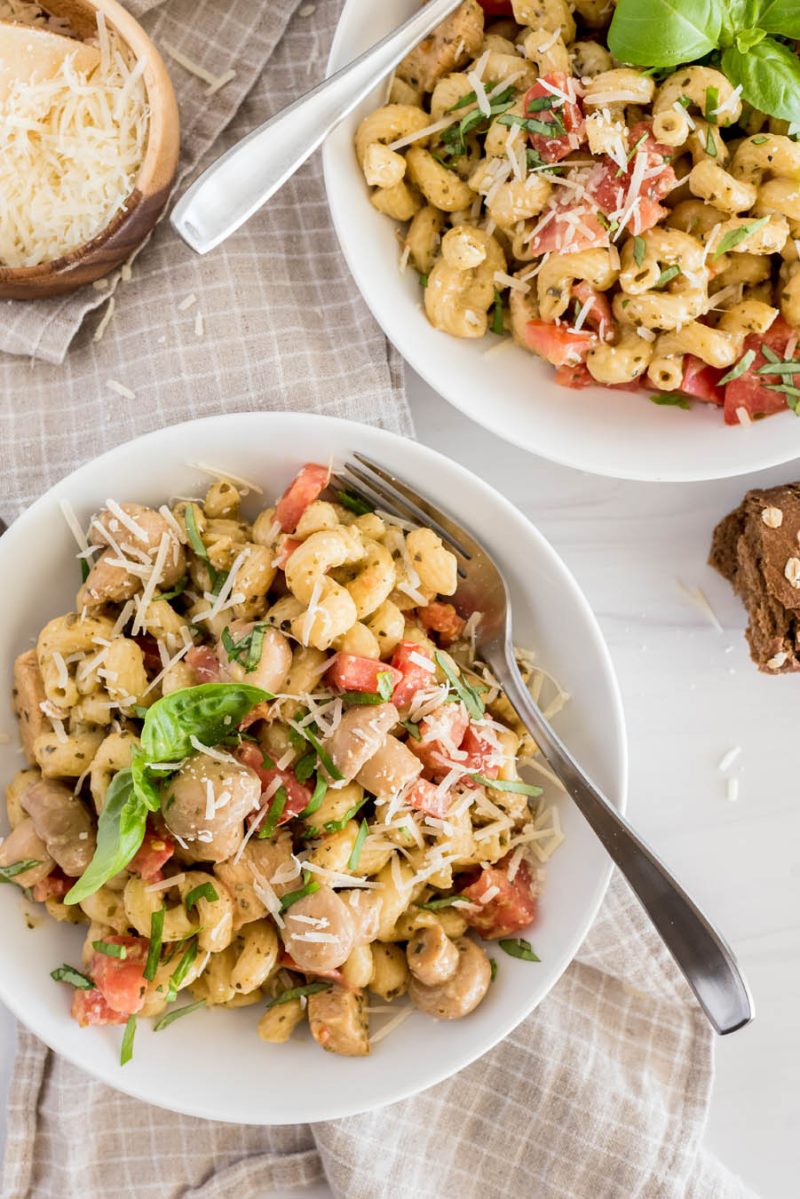 Over head shot of a white bowl with Instant Pot chicken pesto pasta topped with fresh basil and parmesan cheese, with bread and more basil in the background.