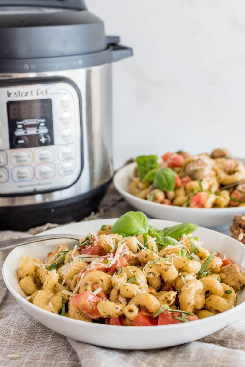 A white bowl filled with pressure cooker chicken pesto pasta topped with fresh basil and shredded parmesan, sitting in front of an Instant Pot.