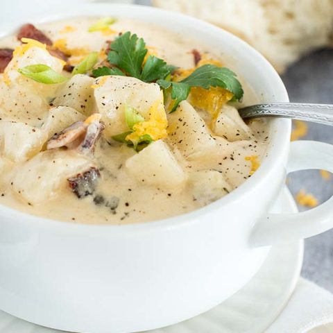 A 45 degree shot of Instant Pot potato soup, made with chunky potatoes, cheddar cheese, and bacon, served in a white soup bowl with a silver spoon tucked in the top right side of the bowl with cheese and bread in the background