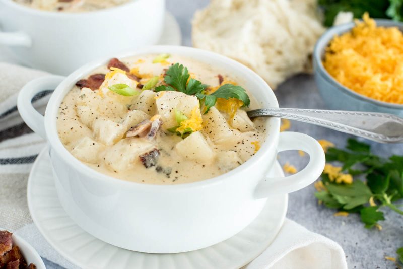 A 45 degree shot of Instant Pot potato soup, made with chunky potatoes, cheddar cheese, and bacon, served in a white soup bowl with a silver spoon tucked in the top right side of the bowl with cheese and bread in the background