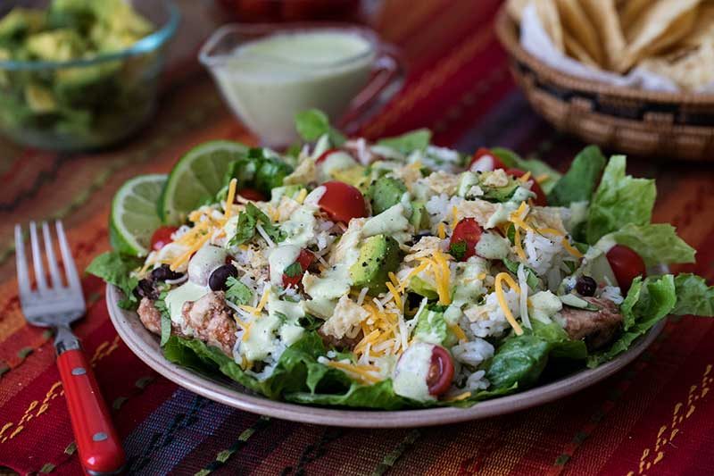 Pressure Cooker Cilantro Lime Chicken Taco Salad shot at a 45 degree angle, with lime slices, dressing, avocado, and chips in the background and a red fork on the side.