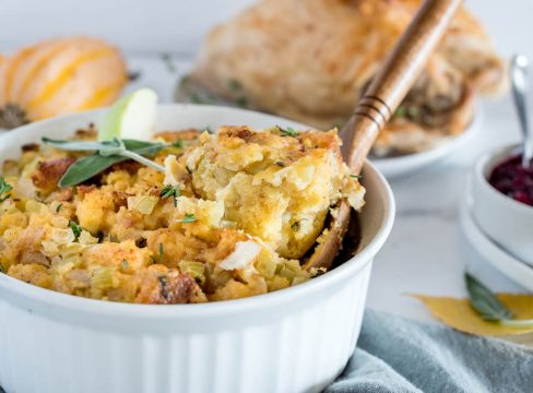 A wooden spoon removing a scoop of Instant Pot cornbread dressing from a white baking dish, with pumpkins and turkey in the background