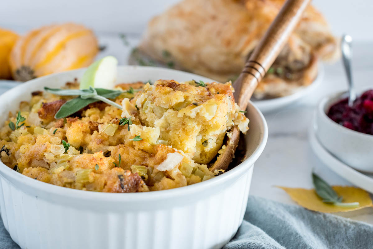 A wooden spoon removing a scoop of Instant Pot cornbread dressing from a white baking dish, with pumpkins and turkey in the background