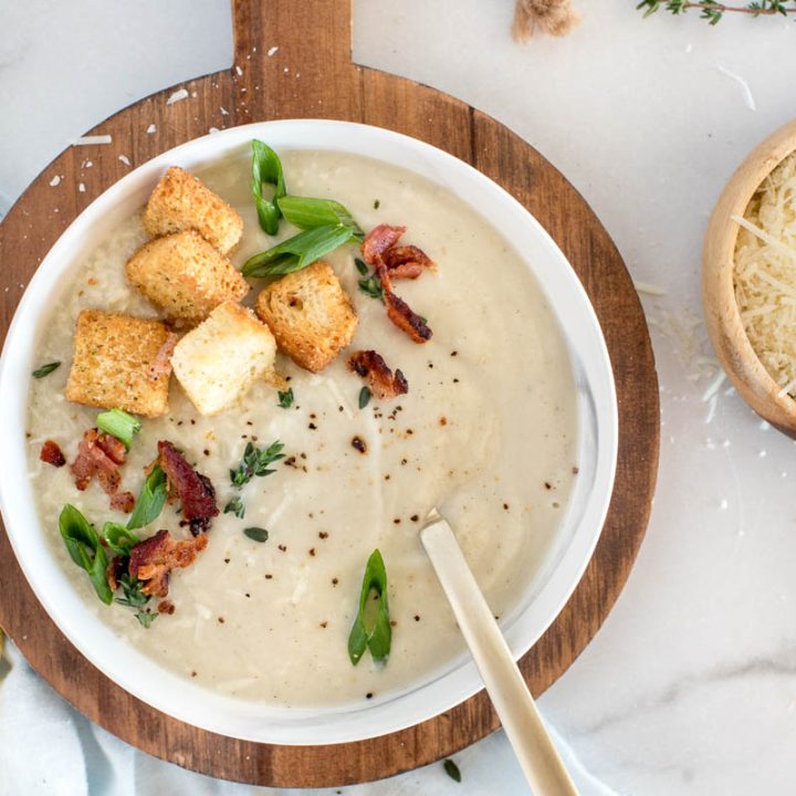 An overhead shot of a white bowl of cauliflower soup topped with croutons, diced green onions, bacon, and extra salt and pepper