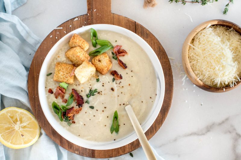 An overhead shot of a white bowl of cauliflower soup topped with croutons, diced green onions, bacon, and extra salt and pepper