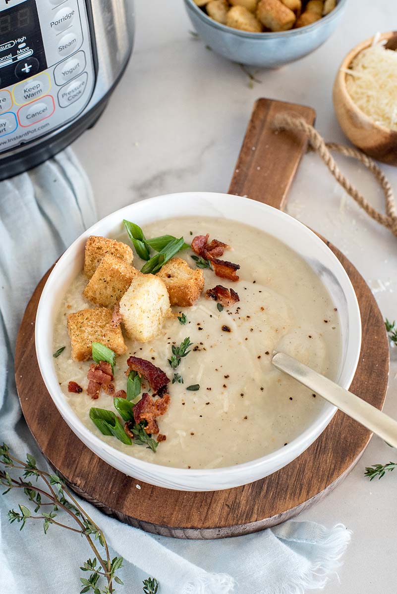 An image of Instant Pot cauliflower soup on a wooden board, with the soup garnished with breadcrumbs , bacon, scallions, and pepper, with an Instant Pot in the background.