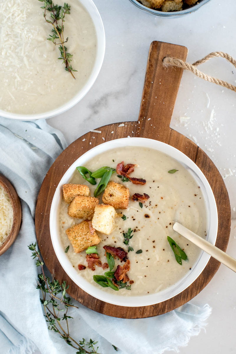 An overhead shot of a white bowl of cauliflower soup topped with croutons, diced green onions, bacon, and extra salt and pepper. Resting on a wooden board, with extra cauliflower soup to garnish at the top