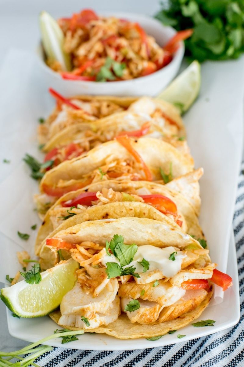 A close-up shot of a fish taco, garnished with lime, bell peppers, and cilantro, with extra tacos in the background