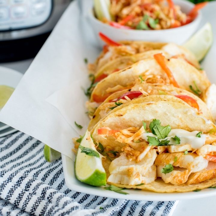 a close-up of a white platter with several fish tacos, garnished with lime, cilantro, fish taco slaw, sitting on a blue and white striped napkin sitting in front of an instant pot