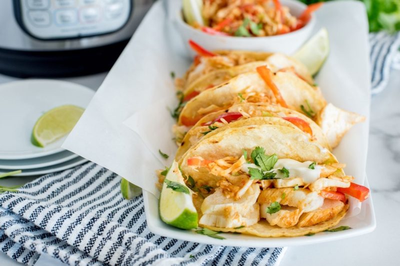 a close-up of a white platter with several fish tacos, garnished with lime, cilantro, fish taco slaw, sitting on a blue and white striped napkin sitting in front of an instant pot