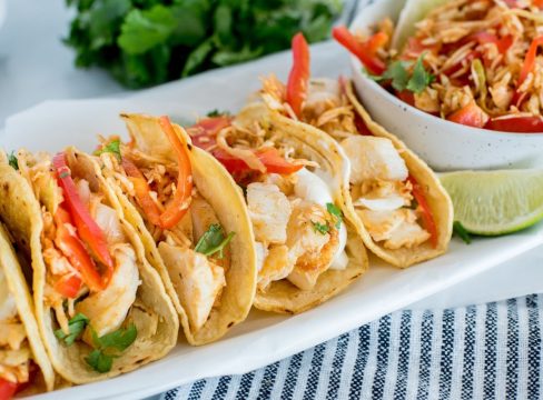 a white platter filled with five fish tacos loaded with cabbage slaw, tomatoes, cilantro, and limes