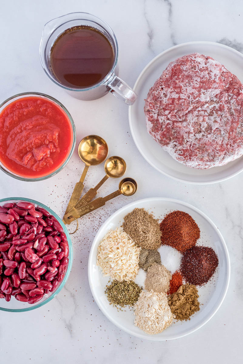 An overhead shot of the ingredients for chili, including frozen ground beef, beef broth, tomato sauce, kidney beans, and a mixture of dried spices