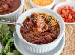 A heaping spoonful of ground beef chili suspended over a small white bowl of ground beef, garnished with shredded cheese, tomatoes, and cilantro