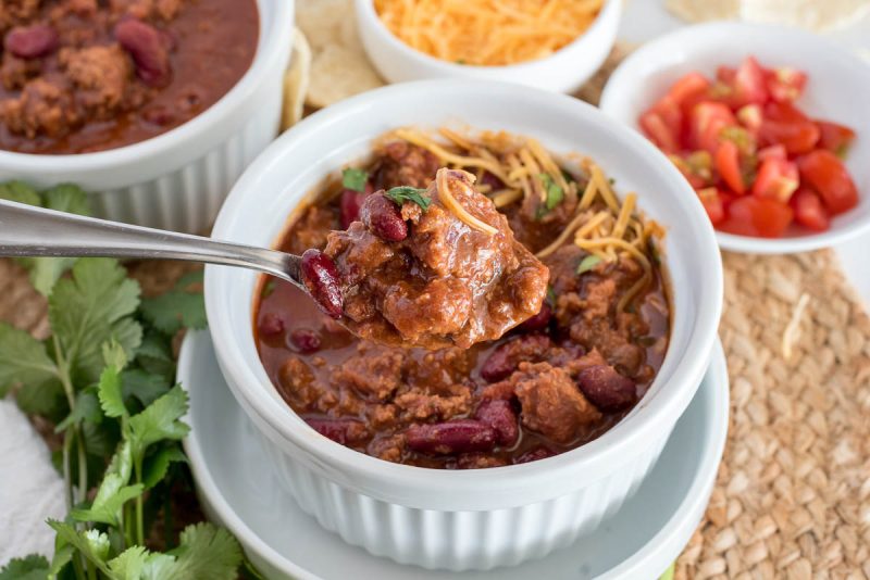 A heaping spoonful of ground beef chili suspended over a small white bowl of ground beef, garnished with shredded cheese, tomatoes, and cilantro