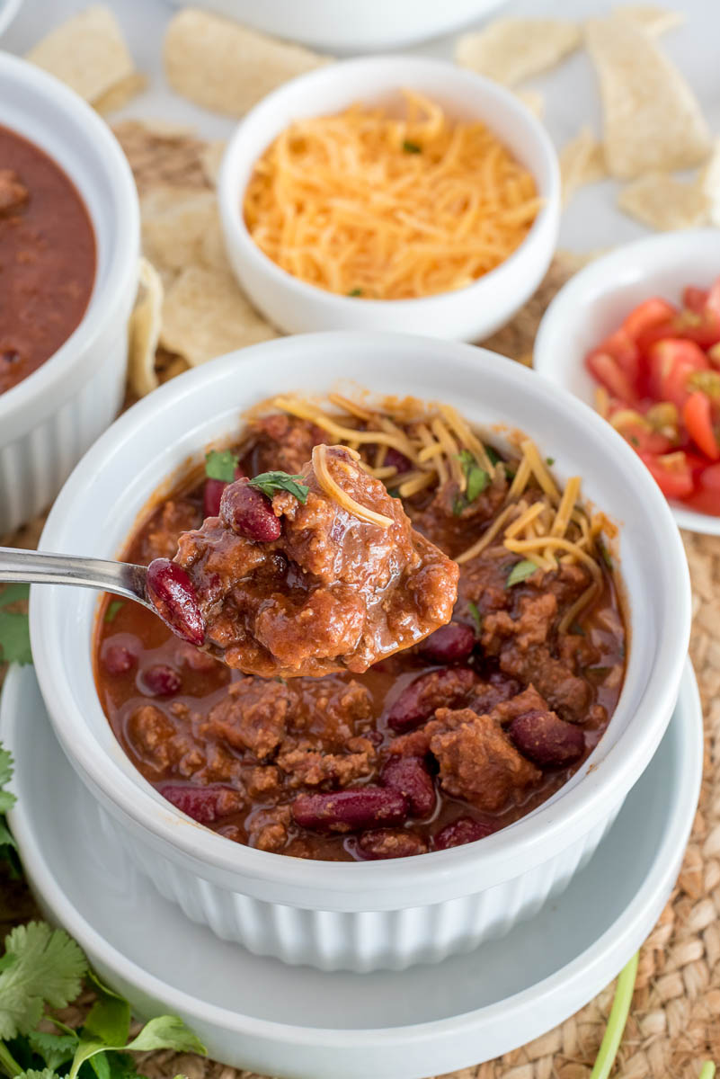 A heaping spoonful of ground beef chili suspended over a small white bowl of ground beef, garnished with shredded cheese, tomatoes, and cilantro