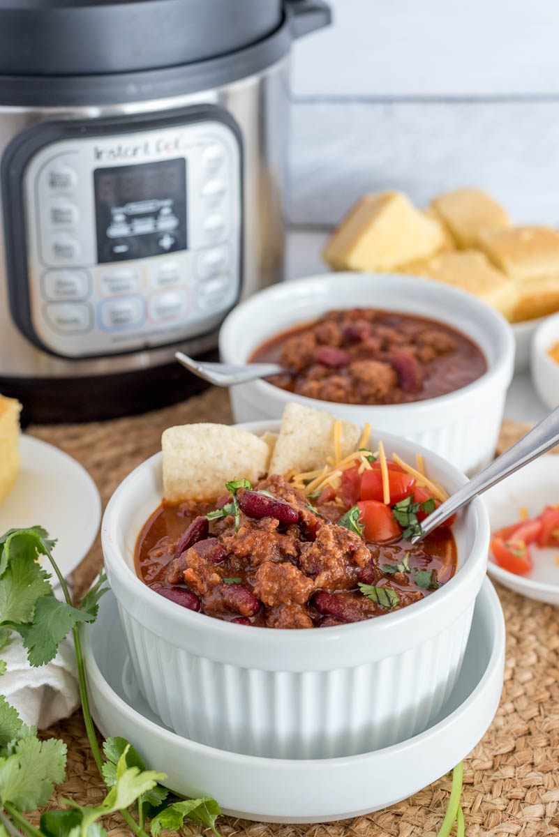 Two small bowls of Instant Pot chili made from frozen ground beef, garnished with diced tomatoes and cilantro and tortilla chips, sitting in front of an Instant pot and a plate of cornbread