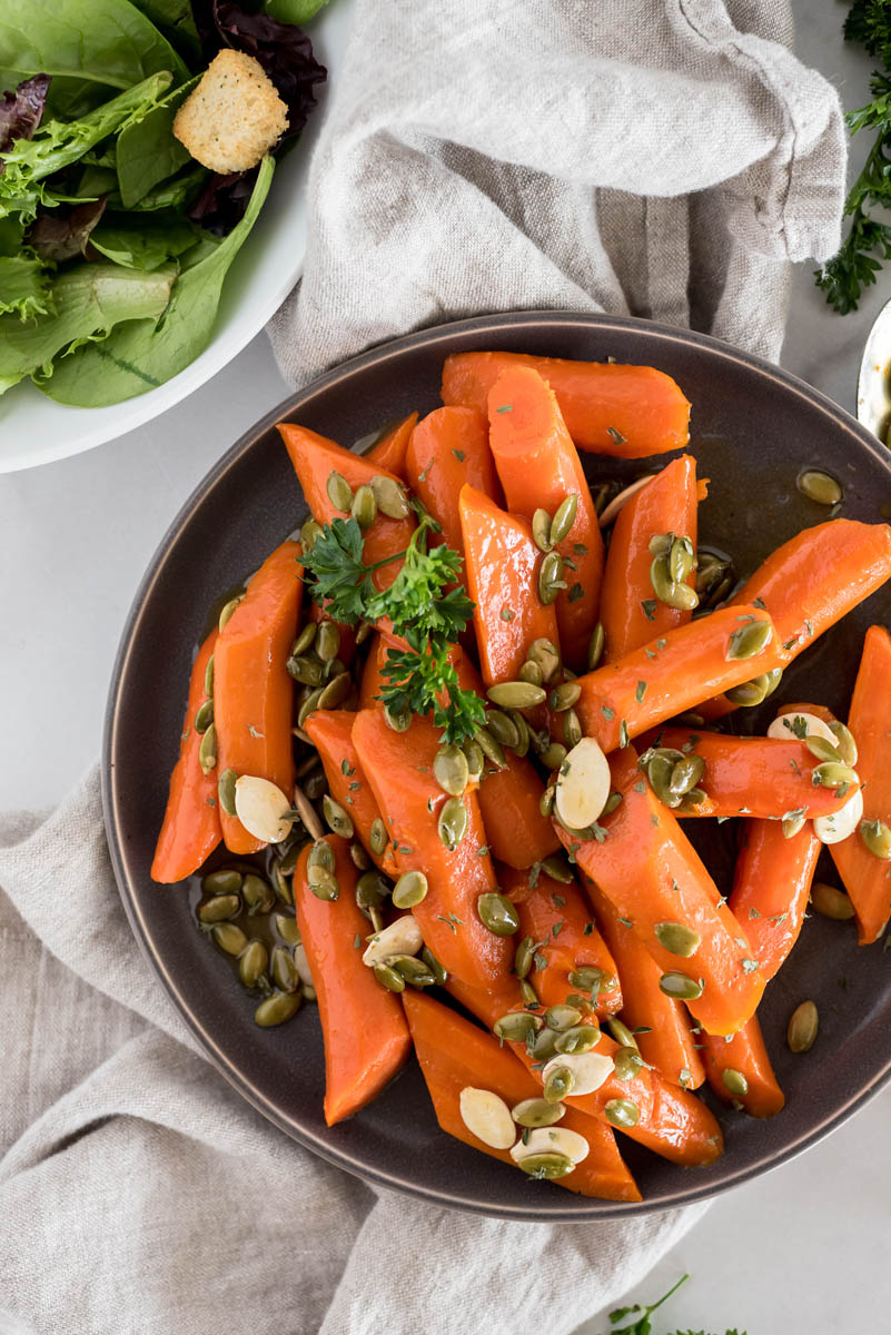 An overhead shot of Instant Pot glazed carrots, with a small dinner salad visible in the top left corner and a beige linen napkin underneath